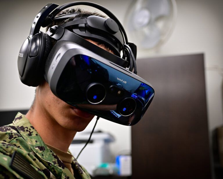 Eine Person in Militärtarnung trägt ein großes Virtual-Reality-Headset und Kopfhörer und steht in einem Innenraum vor einem unscharfen Hintergrund, der einen Ventilator und Bürogeräte zeigt.