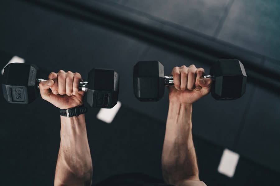 Man sieht die Arme einer Person, die in einem Fitnessstudio zwei schwarze Hanteln hebt. Die Ansicht ist von oben und zeigt die Unterarme und Hände, die die Gewichte halten, mit einem dunklen Hallenboden im Hintergrund.